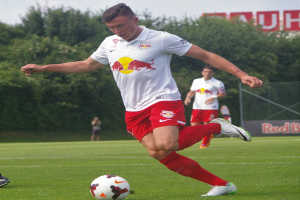Ein professioneller Fussballspieler, der einen Ball auf einem grünen Feld mit Bäumen und einem klaren blauen Himmel im Hintergrund kickt, mit einer Tafel mit der Aufschrift «RB Leipzig» im Hintergrund.