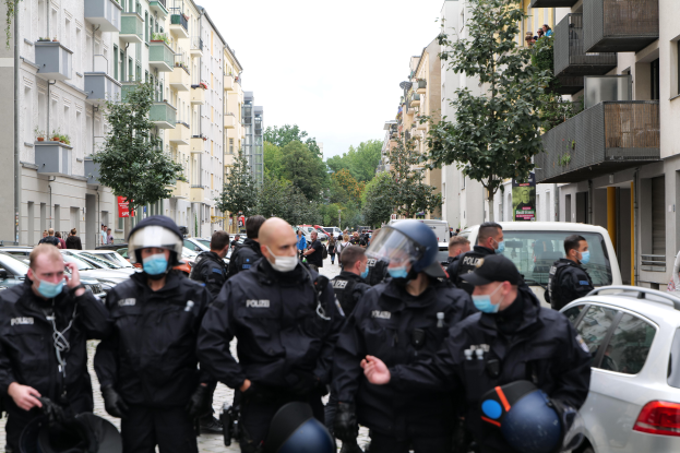 Eine Gruppe von Polizisten in Masken und Helmen marschiert eine Straße mit parkenden Autos entlang, flankiert von Gebäuden mit Balkonen und Geländern, mit Bäumen und einem klaren Himmel im Hintergrund.