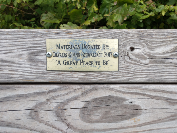 Eine hölzerne Bank mit einer Plakette, auf der "Materialien gespendet von Charles & Ann Schwabach 2017: Ein schöner Ort" steht, vor üppigen grünen Pflanzen.