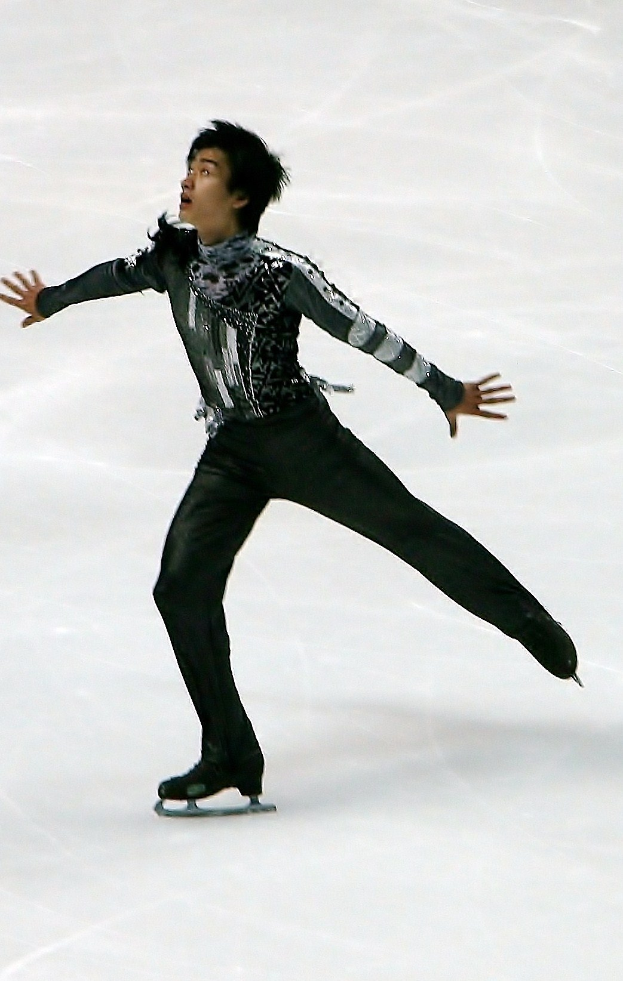 Männlicher Eiskunstläufer in einem schwarzen Kleid, der eine Routine auf einer Eisbahn durchführt und dabei mit synchronen Arm- und Beinbewegungen gleitet.