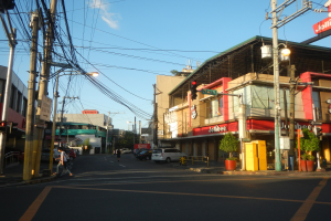 Eine Stadtstraßenszene mit Autos, Fußgängern, Strommasten, Laternen, Ampeln, Gebäuden mit Namensschildern, Blumentöpfen und einem bewölkten Himmel.