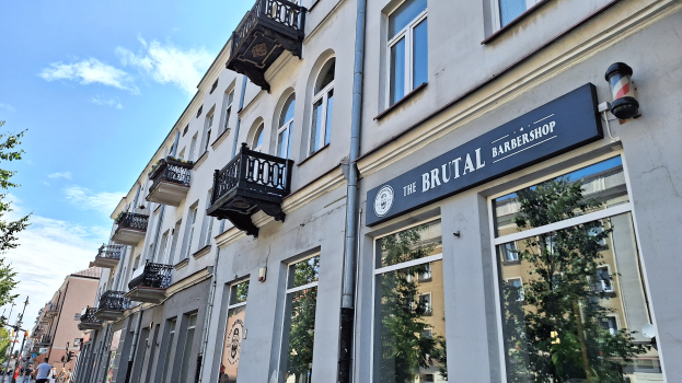 Brutalistischer Barbershop-Laden in Berlin mit Betongebäuden, Fenstern, Geländern, Balkonen und Schildern, Bäumen, Straßenlaternen, Passanten und einer bewölkten Himmel.