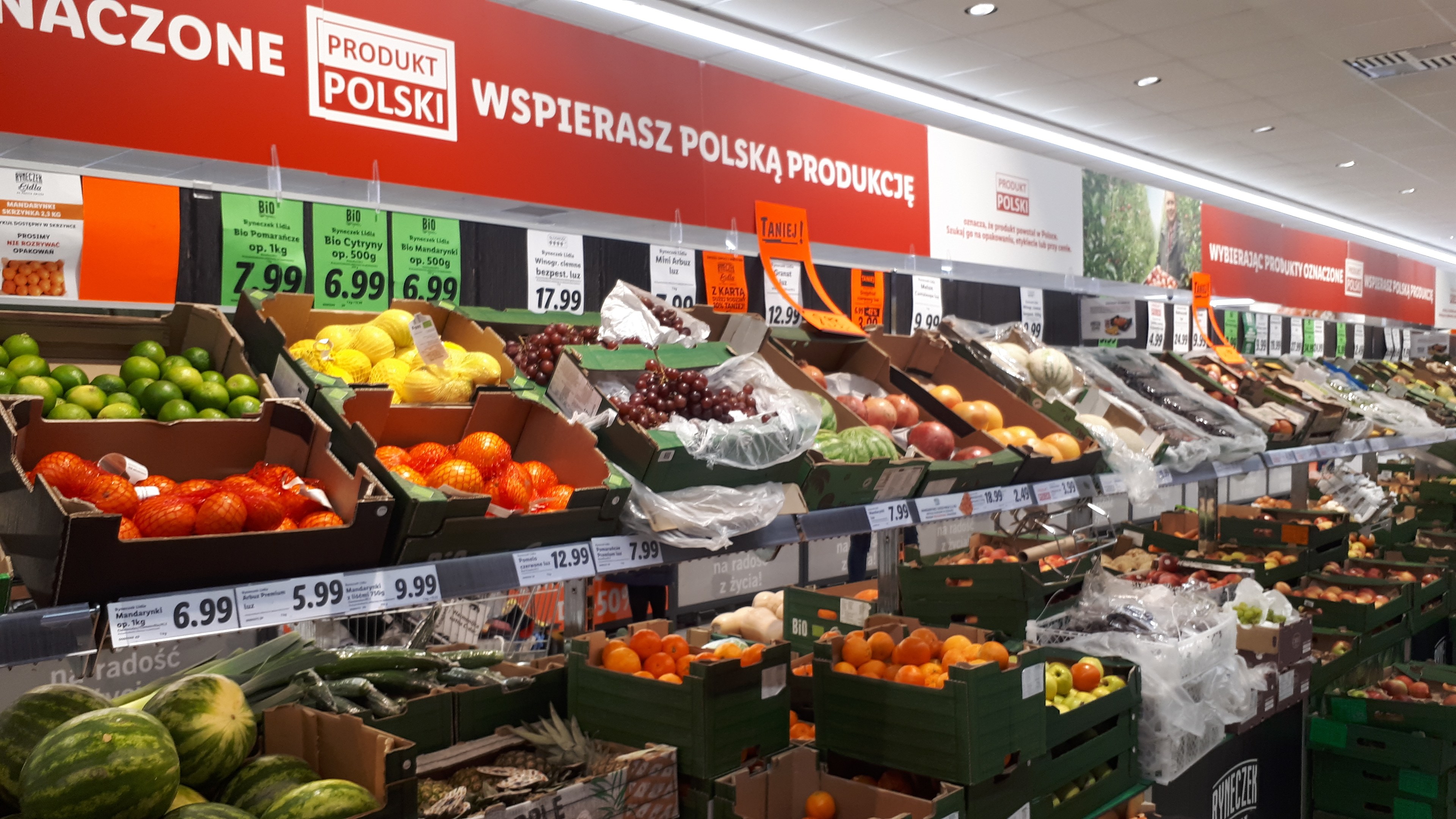 Ladeninneres mit verschiedenen Obst- und Gemüsesorten in Kartonschachteln mit Preisschildern, Werbetafeln an den Wänden und Deckenbeleuchtung.