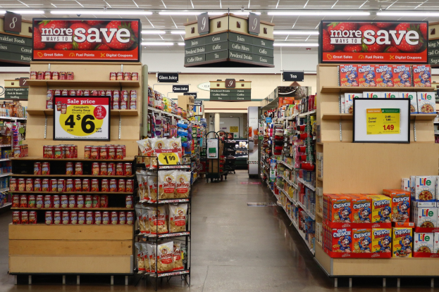 Ein Kiosk-Innenraum mit Regalen und Boden voller verschiedener Lebensmittel, Deckenbeleuchtung, Informationsschilder und einer Person im Hintergrund.