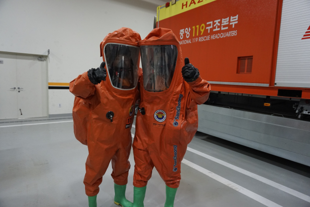 Zwei Personen in orangen Schutzanzögen stehen nebeneinander vor einem Lastwagen, mit einer Wand und Türen im Hintergrund.