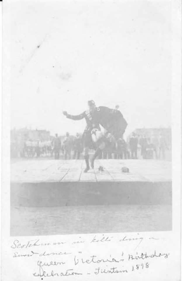Schwarz-weißes Foto eines Mannes in einem Kilt, der auf einer Bühne tanzt, während der Geburtstagsfeier von Queen Victoria in Edinburgh 1898, mit einer Menge und Gebäuden im Hintergrund.