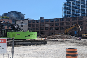 Eine Baustelle mit einem großen grünen Bagger in der Mitte, umgeben von einem Metallzaun und einem Schild an einem Pfahl, mit Maschinen, einem Eimer und einem Verkehrskegel im Vordergrund und Gebäuden, Bäumen und Himmel im Hintergrund.