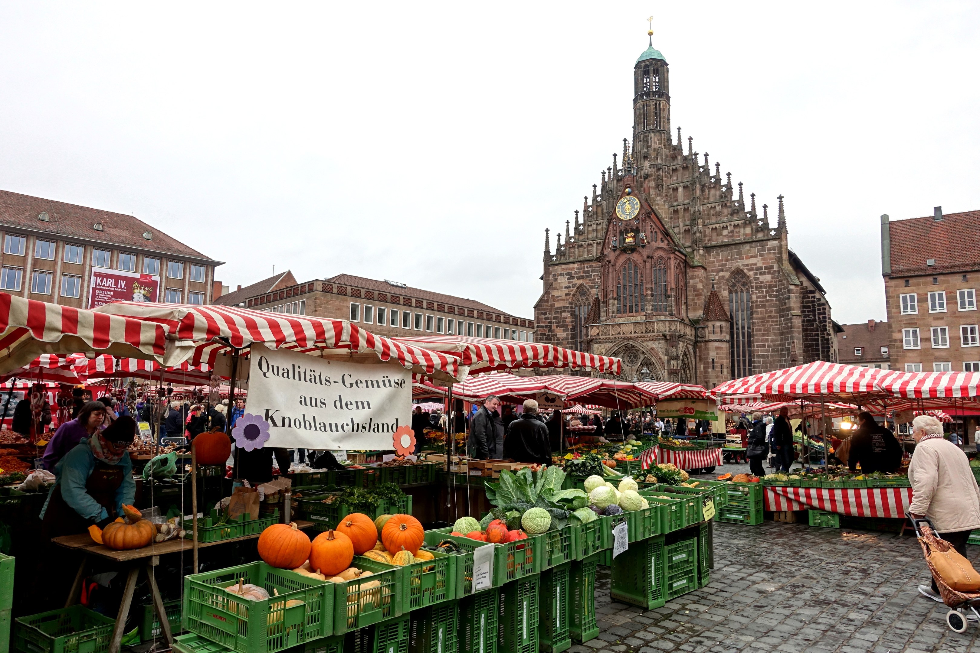 Ein belebter Markt in Nürnberg, Deutschland, mit Obst, Gemüse, Menschen, Zelten, Gebäuden, einem Uhrenturm und Himmel.