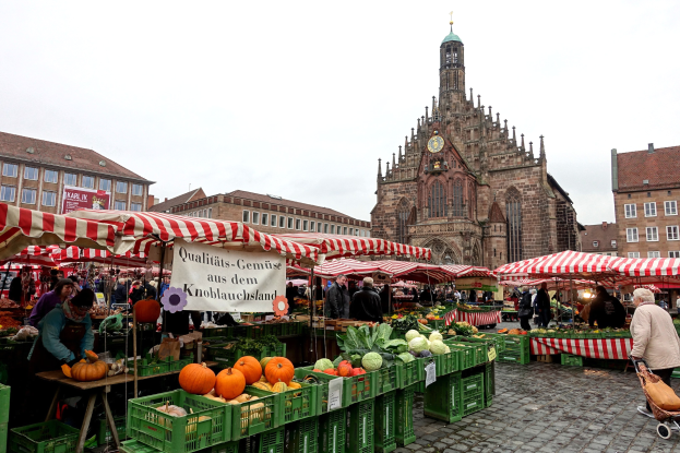 Ein belebter Markt in Nürnberg, Deutschland, mit Obst, Gemüse, Menschen, Zelten, Gebäuden, einem Uhrenturm und Himmel.