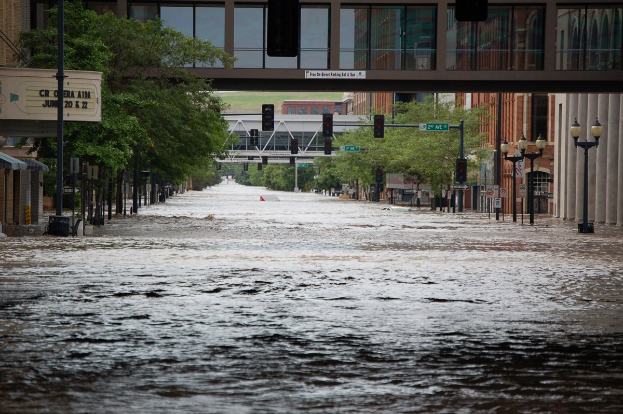 Eine überflutete Stadtstraße mit Wasser, das die Straße, die Infrastruktur und die umliegenden Gebäude bedeckt, mit einer Brücke im Hintergrund.