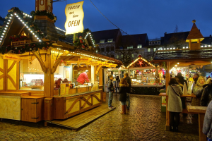 Ein lebendiger Weihnachtsmarkt auf einer Kopfsteinpflasterstraße bei Nacht mit Menschen, die an Ständen mit Lichtern und festlichen Dekorationen vorbeischlendern, Gebäude, Bäume und einen klaren Nachthimmel im Hintergrund.