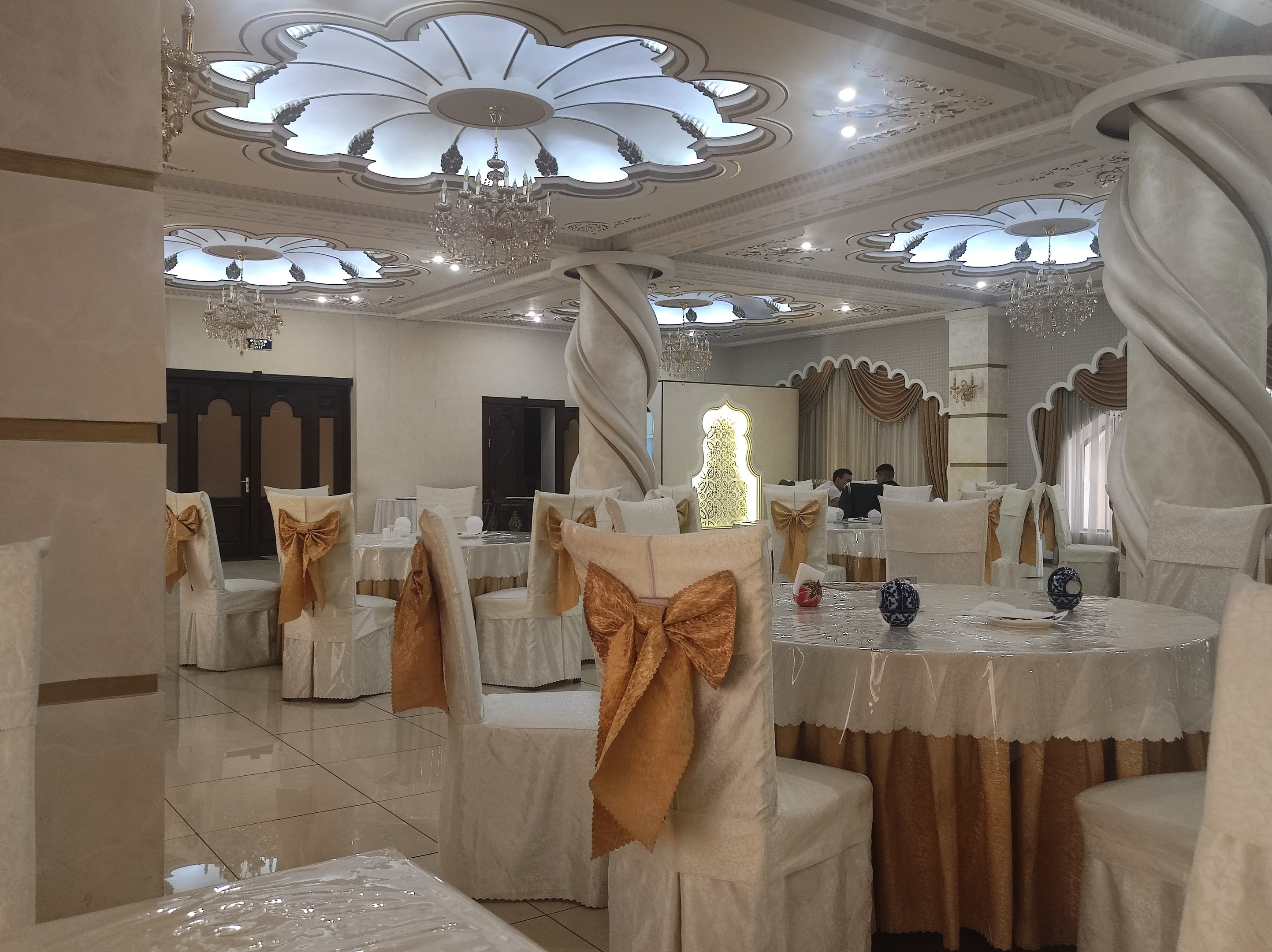 Ein großer Festsaal mit Tischen und Stühlen, die mit Tüchern bedeckt und mit dekorativen Gegenständen geschmückt sind, mit Türen und Vorhängen an den Wänden und beleuchteten Kronleuchtern an der Decke.