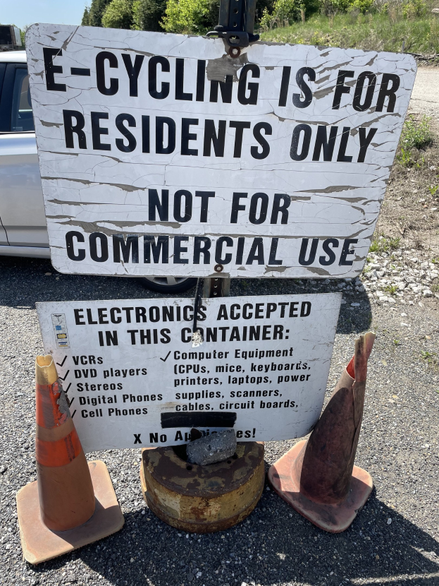 Schild mit der Aufschrift "e-Bike-Fahren nur für Anwohner" neben einer Straße, umgeben von Verkehrskegeln und anderen Gegenständen, mit einem Fahrzeug, Bäumen und einem klaren blauen Himmel im Hintergrund.