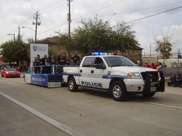 Ein weißer und blauer Polizei-Lkw fährt eine Straße entlang mit einer Gruppe von Menschen auf der Ladefläche, mit Bäumen, Polen, Drähten, Gebäuden und einem klaren blauen Himmel im Hintergrund und einem Geländer mit ein paar Menschen auf dem Gehweg auf der rechten Seite.
