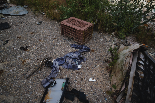 Ein Haufen Müll, einschließlich einer Plastikbox, Kleidung und anderen Gegenständen, die auf dem Boden neben einem Zaun verstreut sind, mit Pflanzen und Gras im Hintergrund.