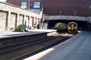 Ein Zug fährt auf den Schienen mit Plattformen auf beiden Seiten, einer Hauswand links, Straßenlaternen, Pflanzen, Bänken, einer Kabine, einer Brücke über die Plattformen und Gebäuden und Masten im Hintergrund.