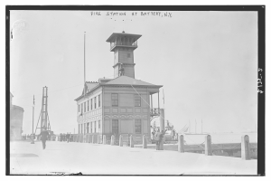 Schwarzes und weißes Foto einer Feuerwache an der Battery, New York, das ein Gebäude mit Fenstern, Menschen, Pfosten, einen Zaun, Boote auf dem Wasser und einen bewölkten Himmel zeigt.