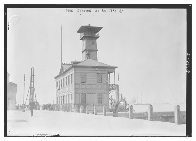 Schwarzes und weißes Foto einer Feuerwache an der Battery, New York, das ein Gebäude mit Fenstern, Menschen, Pfosten, einen Zaun, Boote auf dem Wasser und einen bewölkten Himmel zeigt.