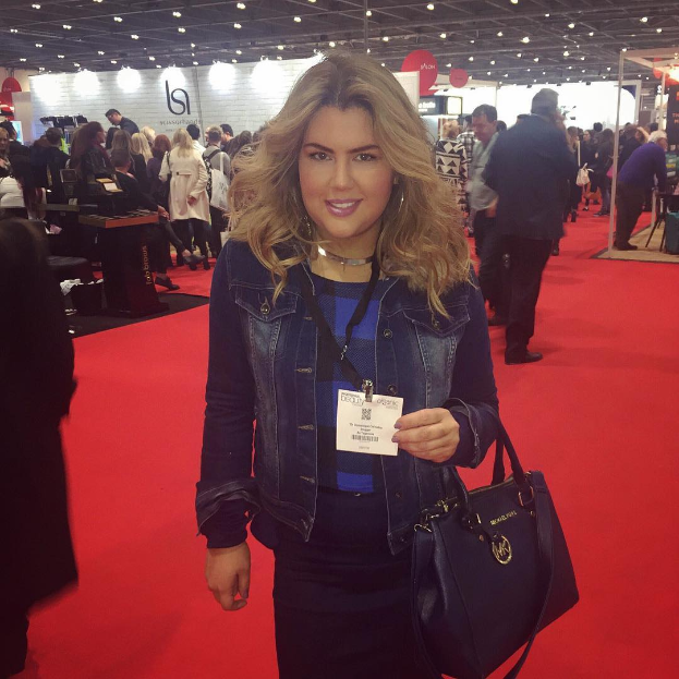 Eine Frau steht auf einem roten Teppich, lächelt und hält eine Handtasche, mit Menschen, Lichtern und Tafeln im Hintergrund bei einer Modenschau.