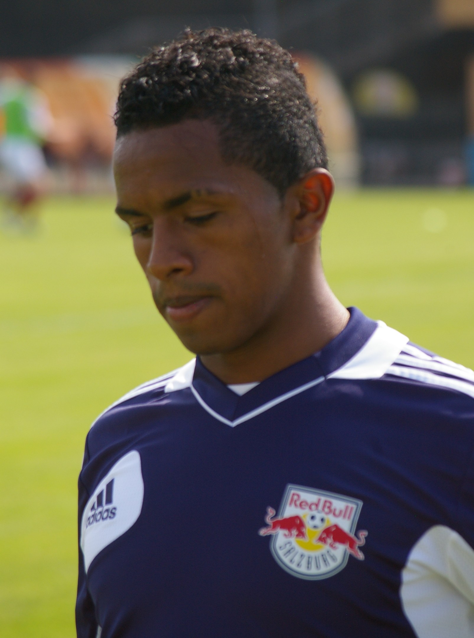 Ein junger Mann in einem blauen T-Shirt steht auf einem Fußballfeld mit entschlossener Miene und sieht aus wie ein Flügelspieler.