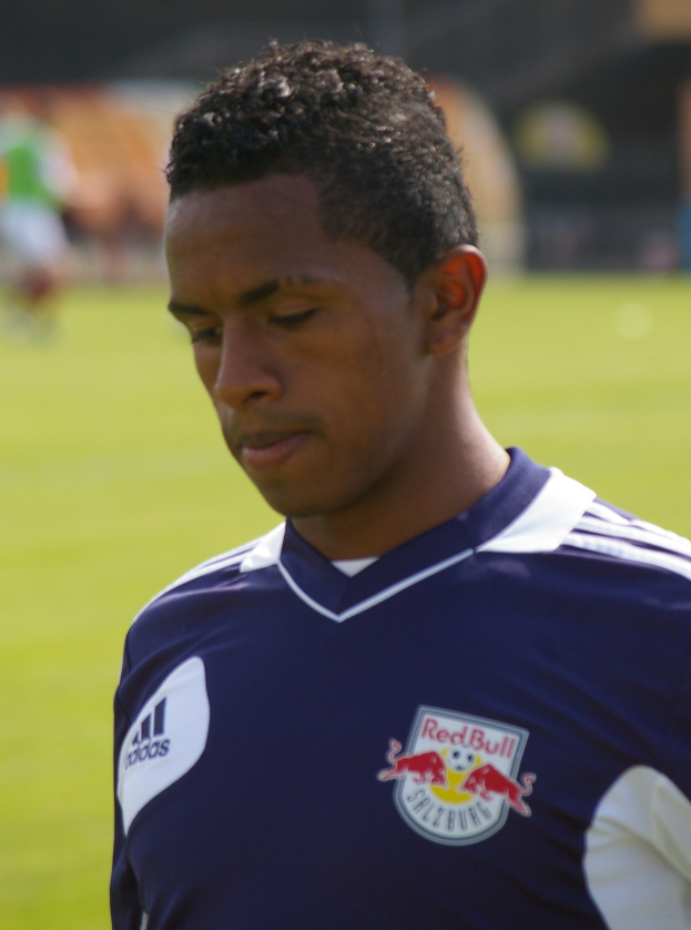 Ein junger Mann in einem blauen T-Shirt steht auf einem Fußballfeld mit entschlossener Miene und sieht aus wie ein Flügelspieler.