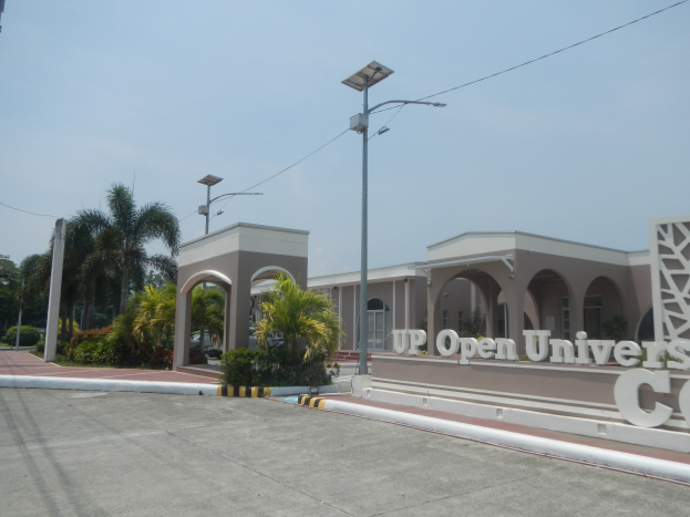Offene Universitätscampus-Eingang mit einer Straße, die zu einem Gebäude führt, umgeben von Bäumen und Pflanzen, eine texttragende Wand auf der rechten Seite und sichtbarer Himmel im Hintergrund.