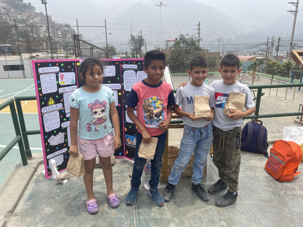 Gruppe von Kindern auf einem Basketballfeld mit Papiertüten in den Händen, mit Gegenständen auf der rechten Seite und einer Tafel mit Text auf der linken Seite, vor einer Hintergrundkulisse aus Zäunen, Pfosten, Straßenlaternen, Bäumen, Gebäuden, Hügeln und Himmel.