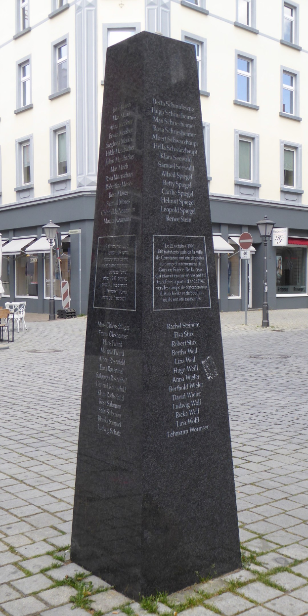 Ein hohes Denkmal mit eingravierter Inschrift steht auf einem Stadtplatz, umgeben von Gebäuden, Straßenmöbeln und Menschen, mit dem Himmel im Hintergrund.