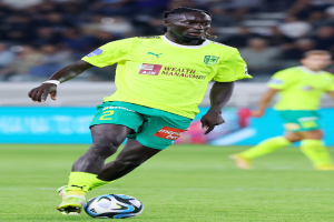 Ein Fußballspieler in einem gelben Trikot und grünen Shorts ist im Einsatz auf dem Feld, mit einer blauen Tafel und unscharfen Figuren im Hintergrund.