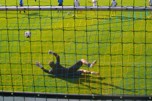 Ein Fussballer taucht ab, um einen Ball vor einem Tor zu halten, mit einem Netz im Vordergrund und einigen Menschen im Hintergrund, während mehrere Fussbälle auf dem Boden verstreut sind.
