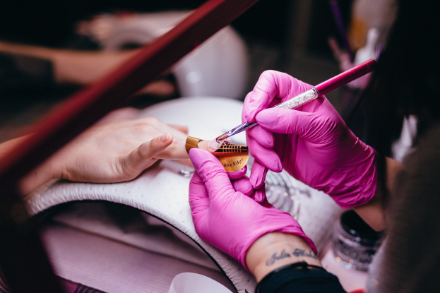 Eine Frau in pinken Handschuhen hält einen Pinsel und eine Nagellackflasche, während sie sich die Nägel in einem Salon machen lässt, mit Maniküre- und Pediküre-Tools auf dem Tisch.