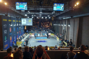 Große Menschenmenge vor einer Bühne mit einem Tischtennis-Tisch in der Mitte, Stühle links, eine Wand rechts und Deckenleuchten und Fernseher im Hintergrund bei der 2019 Jumbo-Weltmeisterschaft.
