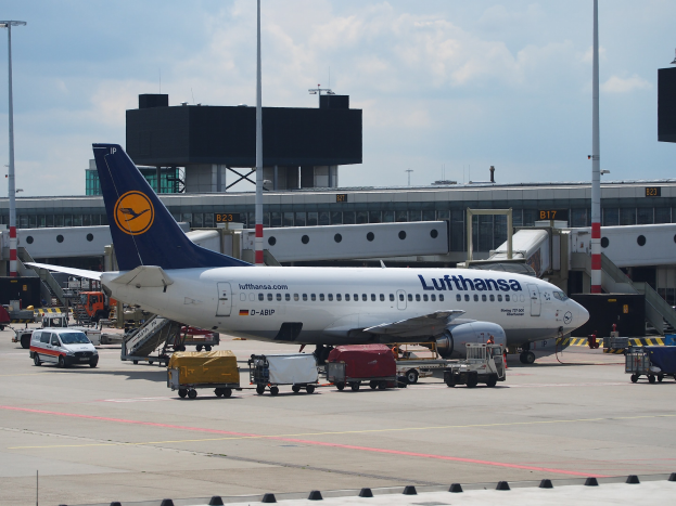 Ein Lufthansa Airbus A320-200-Flugzeug am Frankfurter Flughafen mit Bodenbetriebsfahrzeugen und Wagen im Vordergrund, einem Geb"ude und Laternenmasten im Hintergrund und einem klaren blauen Himmel.