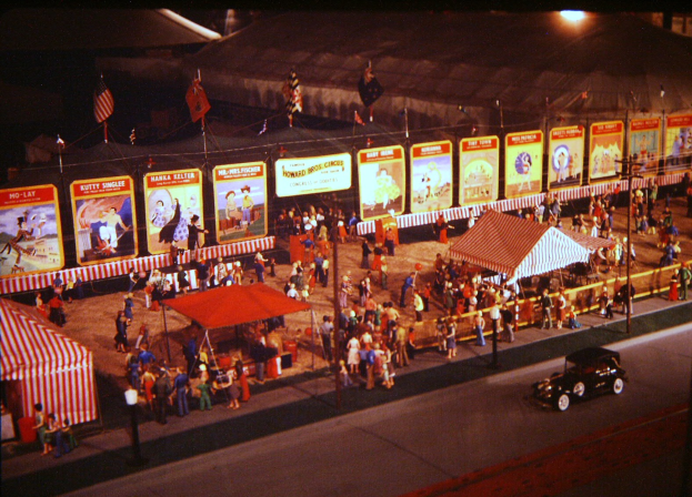 Ein altes Nacht-Karnevalsszenario mit Menschen, einem Auto, Masten mit Fahnen, einem Zaun, einem Zelt, Brettern mit Bildern und Text sowie einer Straßenlaterne.