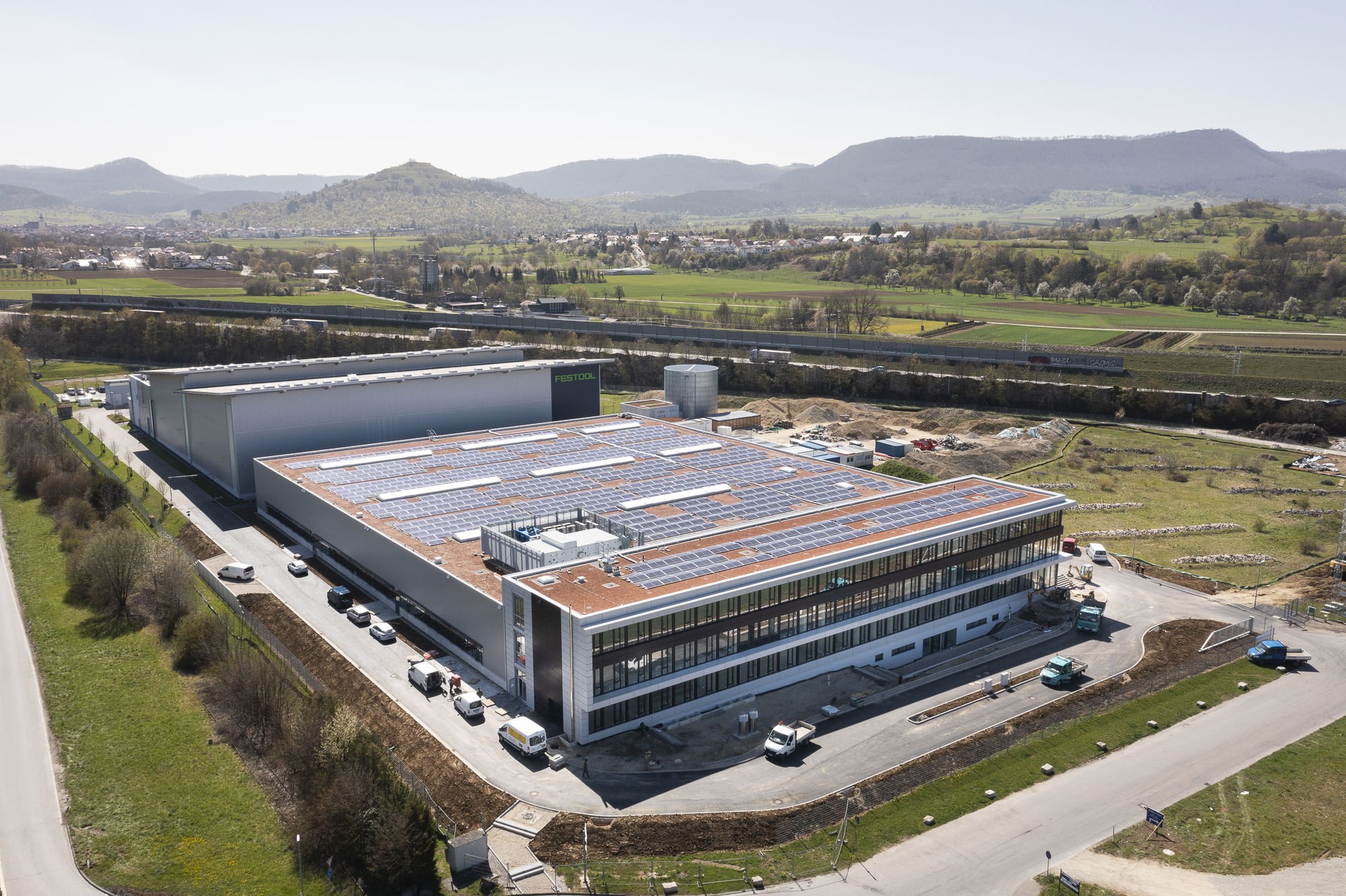 Luftaufnahme eines große Fabriksgebäudes mit Solarpaneelen auf dem Dach, umgeben von Fahrzeugen, Bäumen, Gras und Strommasten, mit Hühgelandschaft und einem klaren blauen Himmel im Hintergrund.