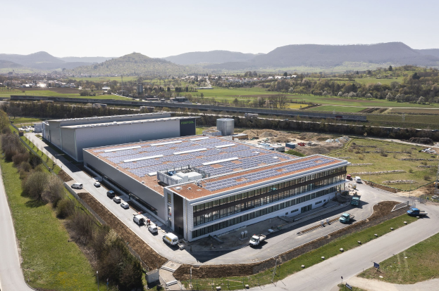 Luftaufnahme eines große Fabriksgebäudes mit Solarpaneelen auf dem Dach, umgeben von Fahrzeugen, Bäumen, Gras und Strommasten, mit Hühgelandschaft und einem klaren blauen Himmel im Hintergrund.
