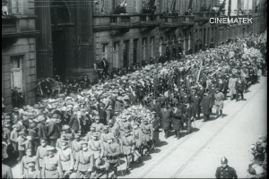 Schwarzes und weißes Foto einer Parade mit einer großen Menge, die eine Straße entlangmarschiert, einige halten Gewehre in den Händen, vor einem Gebäude mit einem Wasserzeichen in der oberen rechten Ecke.