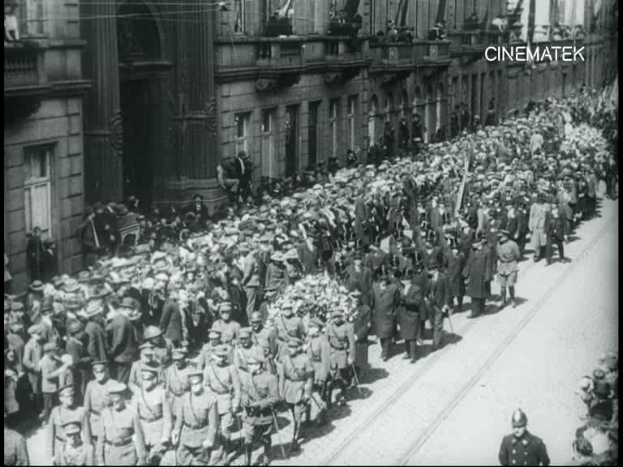 Schwarzes und weißes Foto einer Parade mit einer großen Menge, die eine Straße entlangmarschiert, einige halten Gewehre in den Händen, vor einem Gebäude mit einem Wasserzeichen in der oberen rechten Ecke.