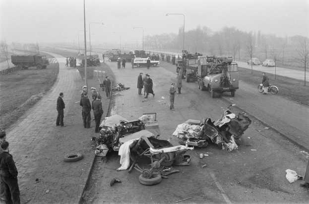 Schwarz-weißes Bild eines Autounfalls am Straßenrand mit mehreren Fahrzeugen und einer Gruppe von Menschen in der Nähe, Lichtmasten, Bäume und Himmel im Hintergrund.