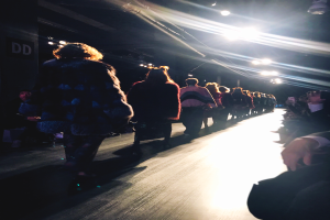 Eine Gruppe von Menschen läuft auf einem hell erleuchteten Laufsteg bei einer Fashion Show, während Zuschauer auf beiden Seiten sitzen.