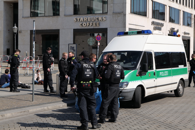 Eine Gruppe von Polizisten steht vor einem Starbucks-Café, rechts steht ein Van und links sitzen und stehen ein paar Menschen, im Hintergrund ist ein Gebäude mit Fenstern, einem Schild, einem Laternenpfahl und einem Zaun zu sehen.