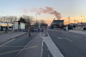 Eine Stadtstraße mit Verkehrskegeln, einem Bus, Laternen, Bäumen, Gebäuden, Kränen und Rauch, der aus einem fernen Gebäude aufsteigt, unter einem klaren Himmel.