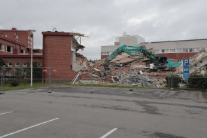 Großes Gebäude in einem Parkplatz mit einem Bagger, Metallstangen, einem Zaun, einem Schild, Pflanzen, Gras, Bäumen, nahen Gebäuden und einem bewölkten Himmel abgerissen.