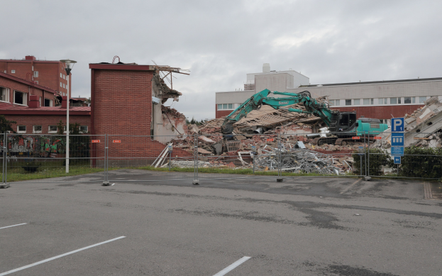 Großes Gebäude in einem Parkplatz mit einem Bagger, Metallstangen, einem Zaun, einem Schild, Pflanzen, Gras, Bäumen, nahen Gebäuden und einem bewölkten Himmel abgerissen.