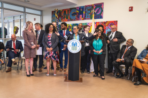 Gruppe von Menschen vor einem Podium mit einem Mikrofon und einer Tafel mit Text, einige sitzen auf Stühlen und andere stehen, mit Fahnen, Feuerlöscher und Deckenlampen an der Rückwand.