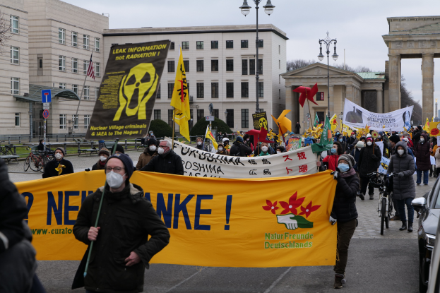 Große Gruppe von Menschen bei einer Straßendemonstration gegen Atomkraft in Deutschland, die Transparente und Fahnen schwenkt, mit Fahrzeugen und Gebäuden im Hintergrund.