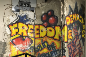 Mauer mit Graffiti, das "Freiheit und Demokratie" in großen schwarzen Buchstaben liest, ein Geländer unten und ein Gebäude mit Fenstern im Hintergrund.