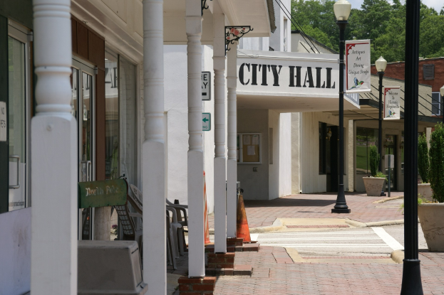 Rathausgebäude mit umliegender Infrastruktur, einschließlich Straßenlaternen, Schildern, Fahrzeugen, Verkehrskegeln, Stühlen, Topfpflanzen, Bäumen und einer bewölkten Himmel.