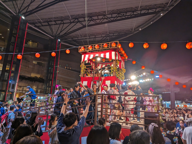 Menschenmenge auf einer beleuchteten Bühne mit Laternen, einige halten Kameras, umgeben von einem Geländer, mit Bäumen und einem blauen Himmel im Hintergrund, möglicherweise während eines japanischen Festivals.