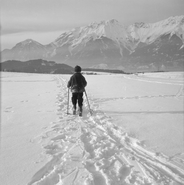 Eine Person, die im Schnee Skilanglauf betreibt, mit Bergen im Hintergrund, die eine Jacke, eine Mütze und Handschuhe trägt und Ski-Stöcke hält.